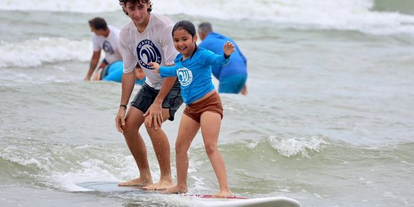 A young girl learning to surf with an instructor on a calm ocean beach.