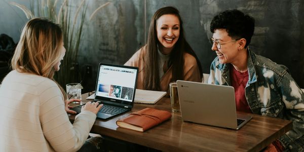 Three friends happily working together on laptops at a wooden table.
