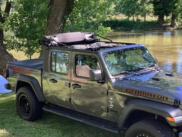 Jeep Gladiator with open soft top parked by a river under trees.