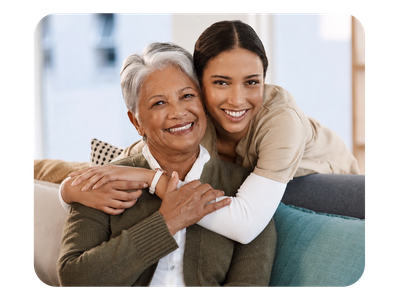 A young woman affectionately hugging an elderly woman on the couch, both smiling warmly.
