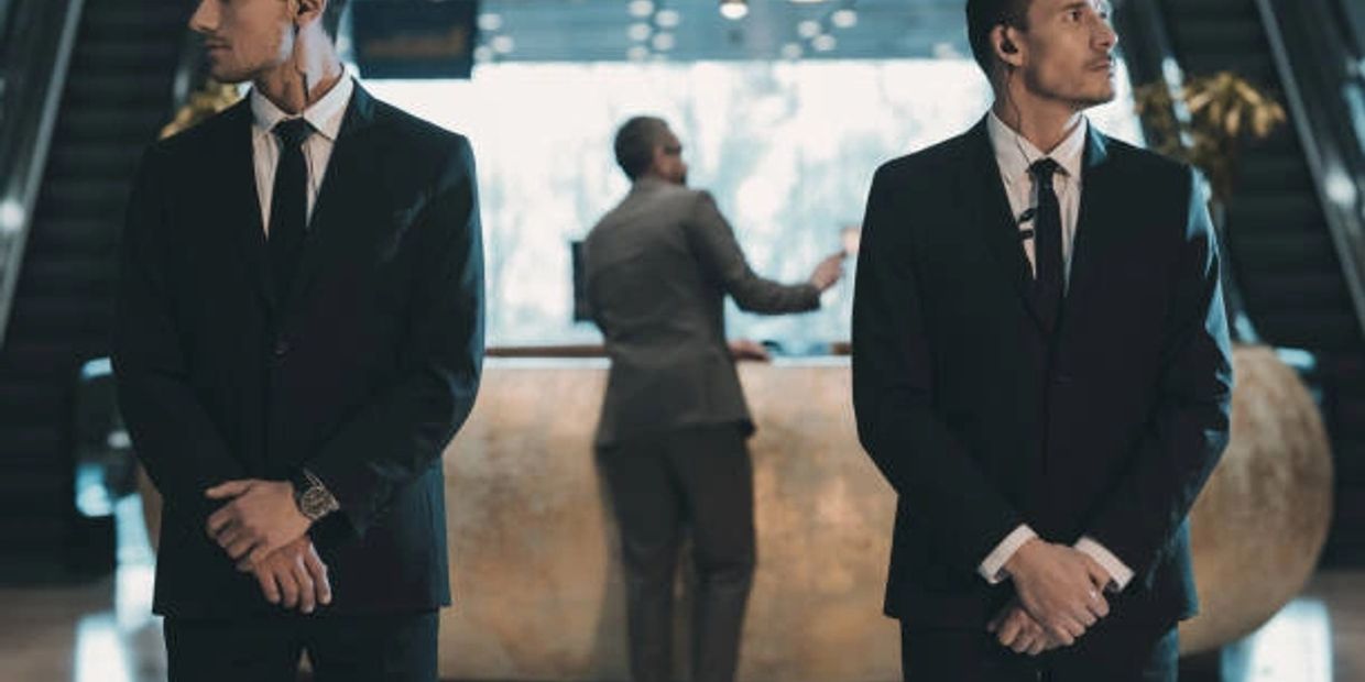 Two security guards in suits stand watch while a man checks in at a hotel reception.