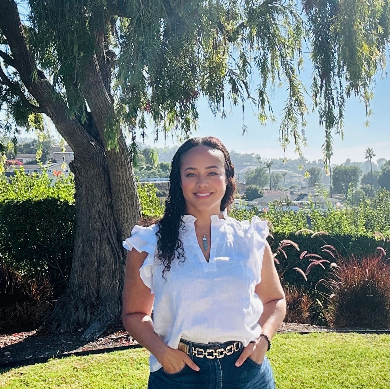 A woman smiling outdoors under a tree on a sunny day.
