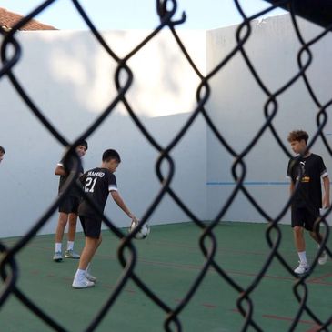 Teen boys playing soccer on a fenced court.