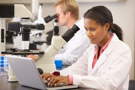 Student in lab coat typing on laptop next to a microscope.