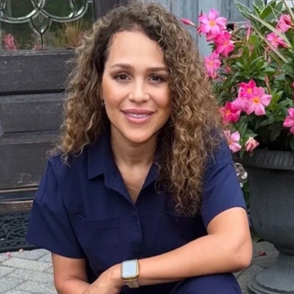 Woman with curly hair smiles in navy blue outfit near pink flowers.
