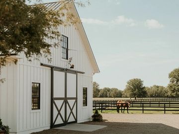 A white barn next to a horse pasture with one horse grazing behind the black fence posts.