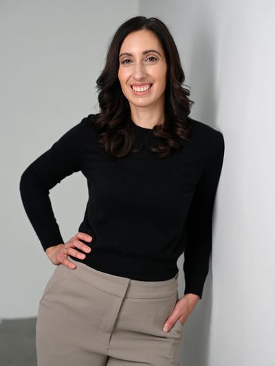 Woman with dark hair smiling, dressed in a black sweater and beige pants.