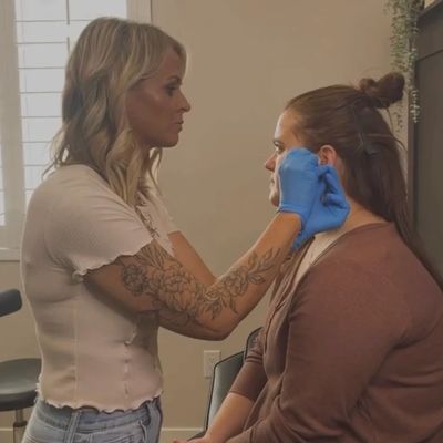 A woman wearing blue gloves is carefully applying makeup on another woman sitting down.