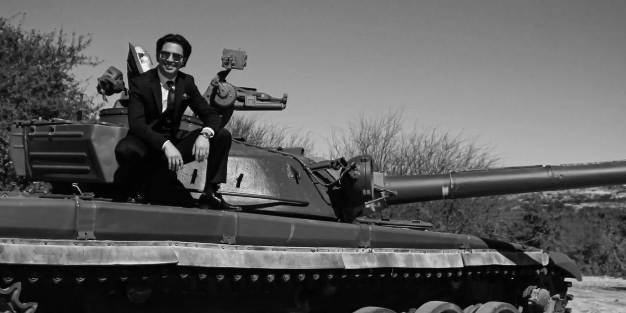A man in a suit sits on a tank, smiling confidently outdoors.