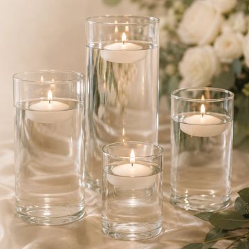 Four glass vases with floating candles and white flowers in background.