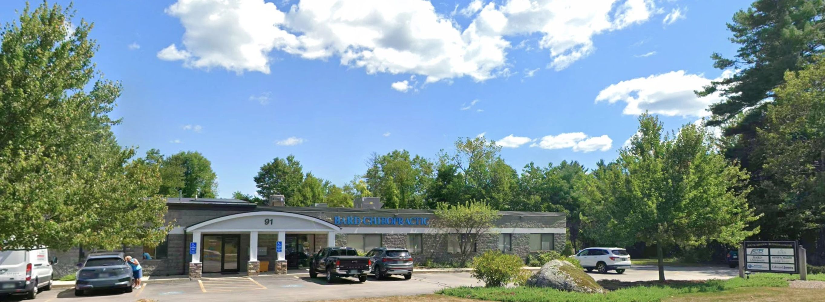 Chiropractic clinic building with parking lot and surrounding trees on a sunny day.