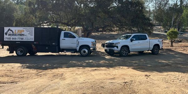 Two white demolition trucks parked on a dirt lot under trees.