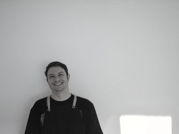 Smiling man in dark clothing against a plain light wall.