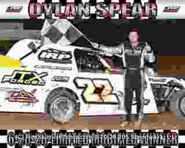 Race car driver Dylan Spear stands next to his winning car holding a checkered flag.