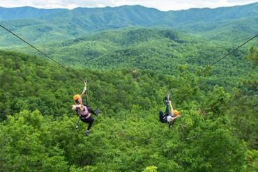 Two people ziplining over lush green forest and mountains.