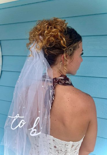 Bride with curly hair wearing a veil with pearls and a brown halter top.
