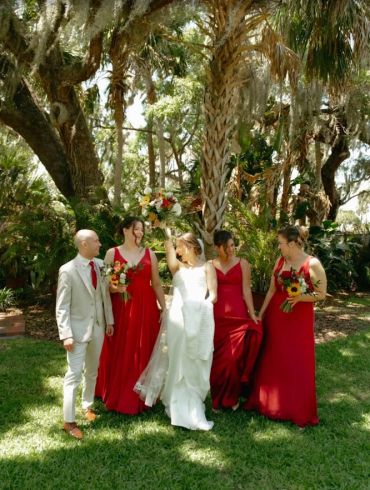 Bride and wedding party outdoors with red dresses and light suit.