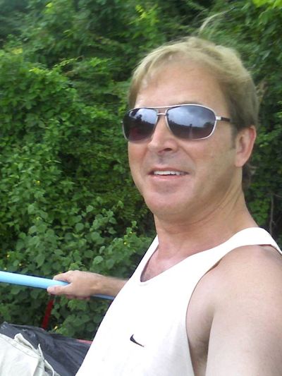 Man wearing sunglasses and a white tank top outdoors near greenery.