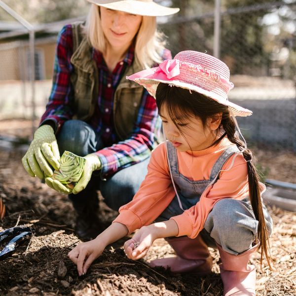 A woman wearing a sun hat looks on while a young child carefully places seeds in a garden furrow.