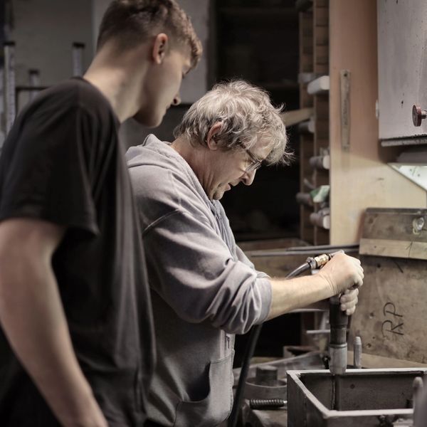 A young man watches an older mentor demonstrate shop work.