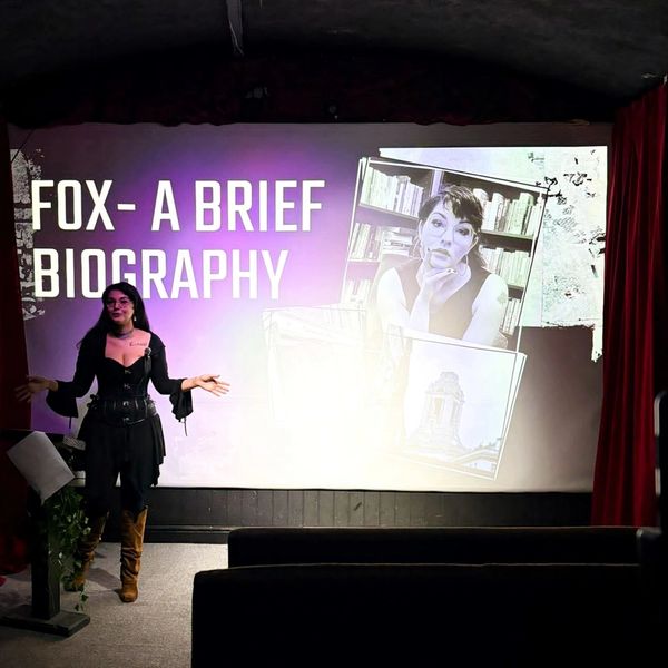 A woman presents a talk titled "FOX- A BRIEF BIOGRAPHY" with a projector screen behind.