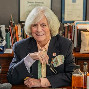 Smiling older woman in suit sitting at a desk with books and drinks.