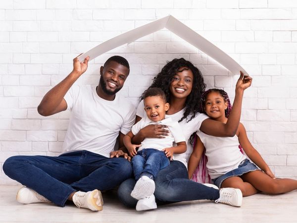 Young African American Parents With Two Children Sitting Under Cartboard Roof Dreaming Of New Home