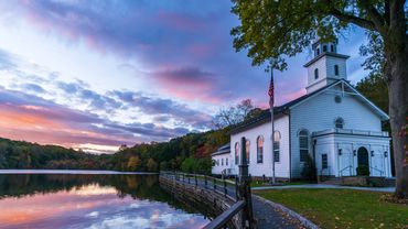 White church beside calm lake at sunset with colorful sky reflection.