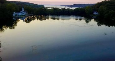 Calm lake reflecting trees and a church at dusk with distant hills.