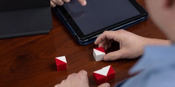 Person assembling a red and white puzzle on a wooden table with a tablet nearby.