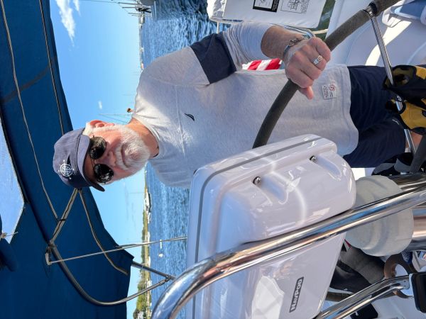 Older man steering a boat on a sunny day by the water.