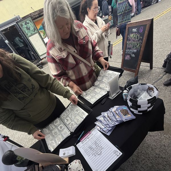Two women browsing tattoo designs at an outdoor market booth.