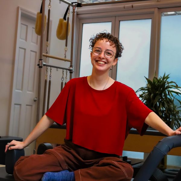 Odessa smiles warmly at the camera. She sits on a grey and white reformer with a red shirt, brown pa