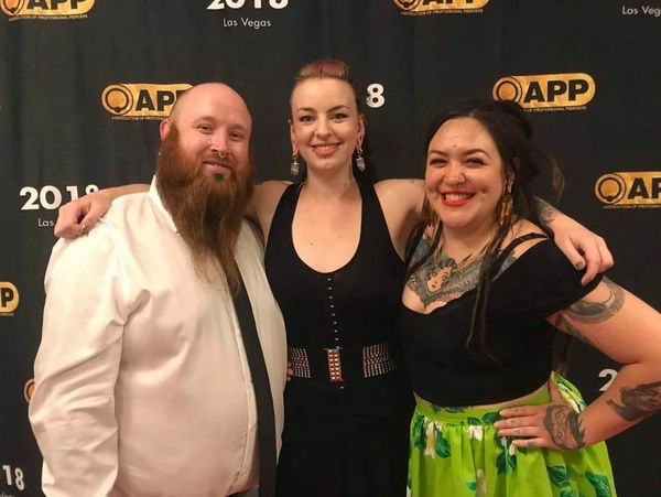 Three tattooed and pierced people embracing and smiling in front of an event backdrop for 'APP 2018'
