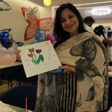 Woman proudly shows her colorful flower drawing in a lively room with balloons and art supplies.