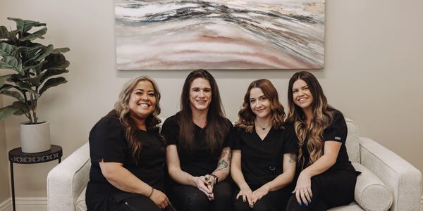 Four women in black scrubs smiling on a white couch in a cozy room.