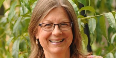 Smiling woman with glasses outdoors near green foliage.