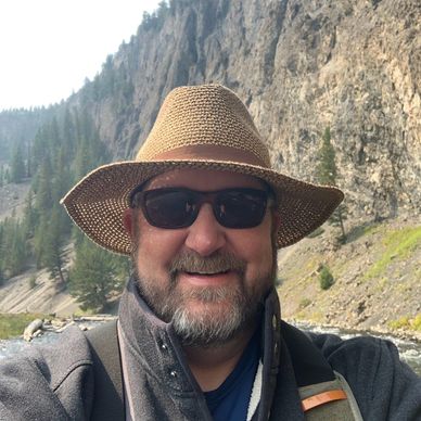 Man wearing a straw hat and sunglasses smiling outdoors by a rocky river.