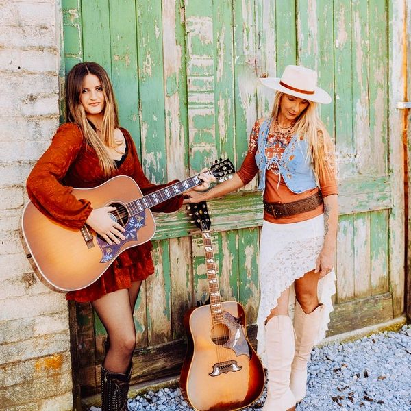 Two women with guitars pose in front of a rustic green wooden door.
