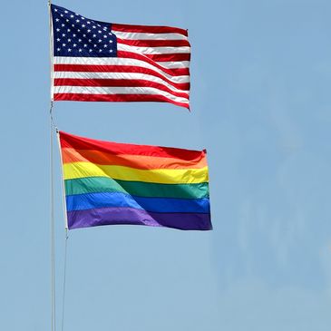 American flag and rainbow pride flag flying on flagpoles against a clear blue sky.