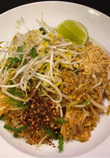 Plate of pad thai with bean sprouts, crushed peanuts, chili flakes, and lime wedge.