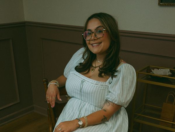 A smiling woman in glasses and a white dress sitting on a chair indoors.
