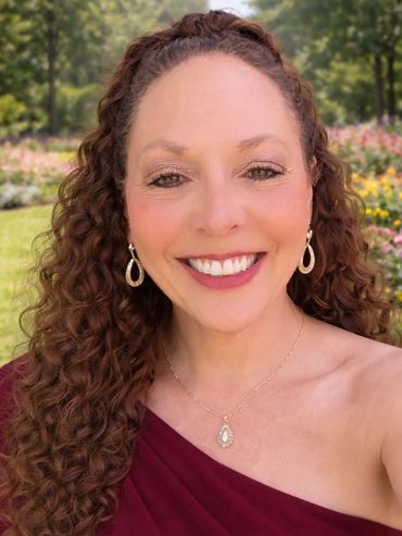 Smiling woman with curly hair in a burgundy dress outdoors.