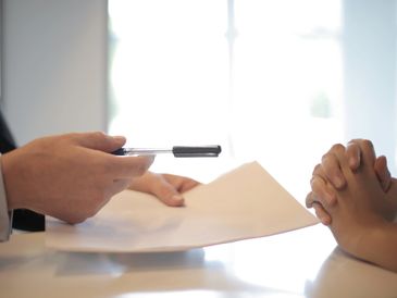 Person holding a pen and paper across from another person with clasped hands.