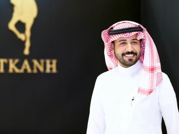 Smiling man in traditional Saudi attire stands beside a black wall with a golden horse logo and the text 'TKANH'.