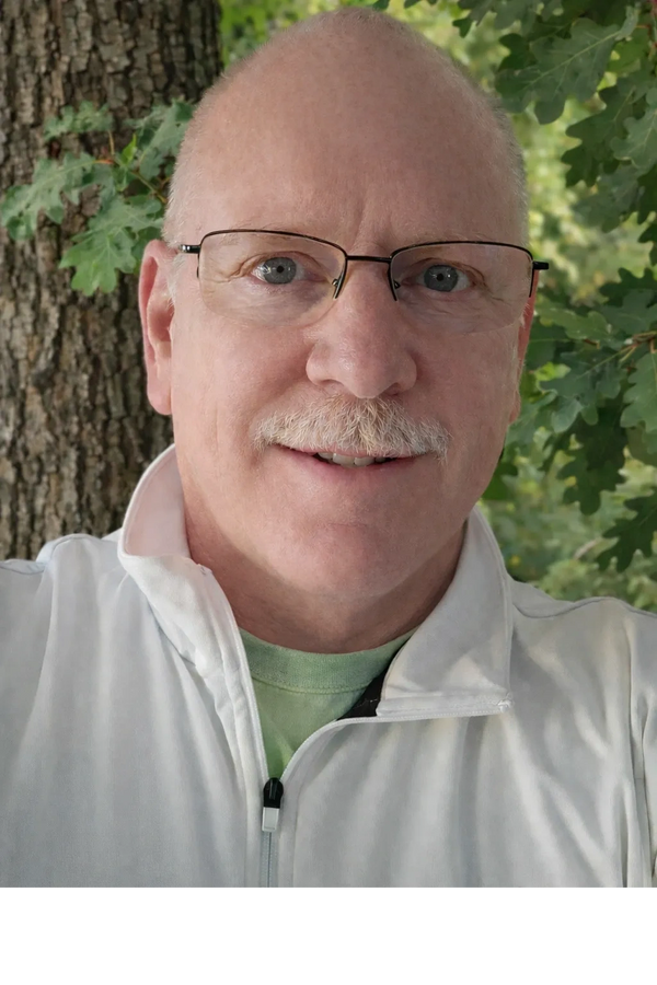 A bald man with glasses and a mustache smiles outdoors near a tree.