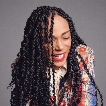 Person with long twisted hair laughing warmly, wearing a colorful patterned jacket.