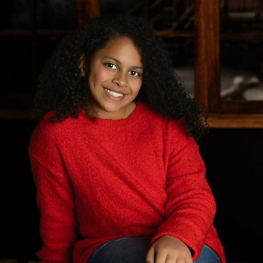 A girl in a red sweater smiles warmly indoors by a window.