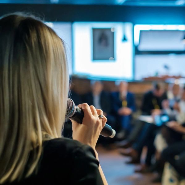 Blond woman speaking into a microphone in front of a seated audience.