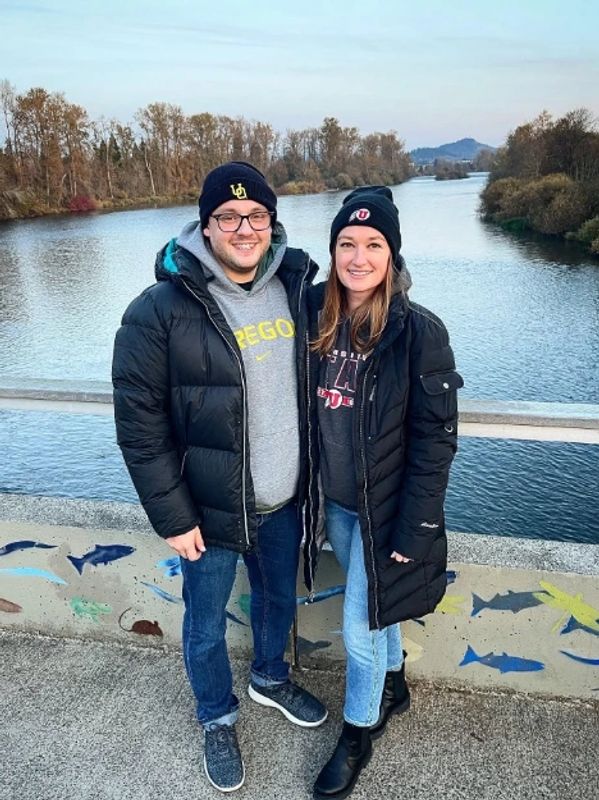 A couple in winter jackets standing by a river on a bridge with painted fish on the railing.